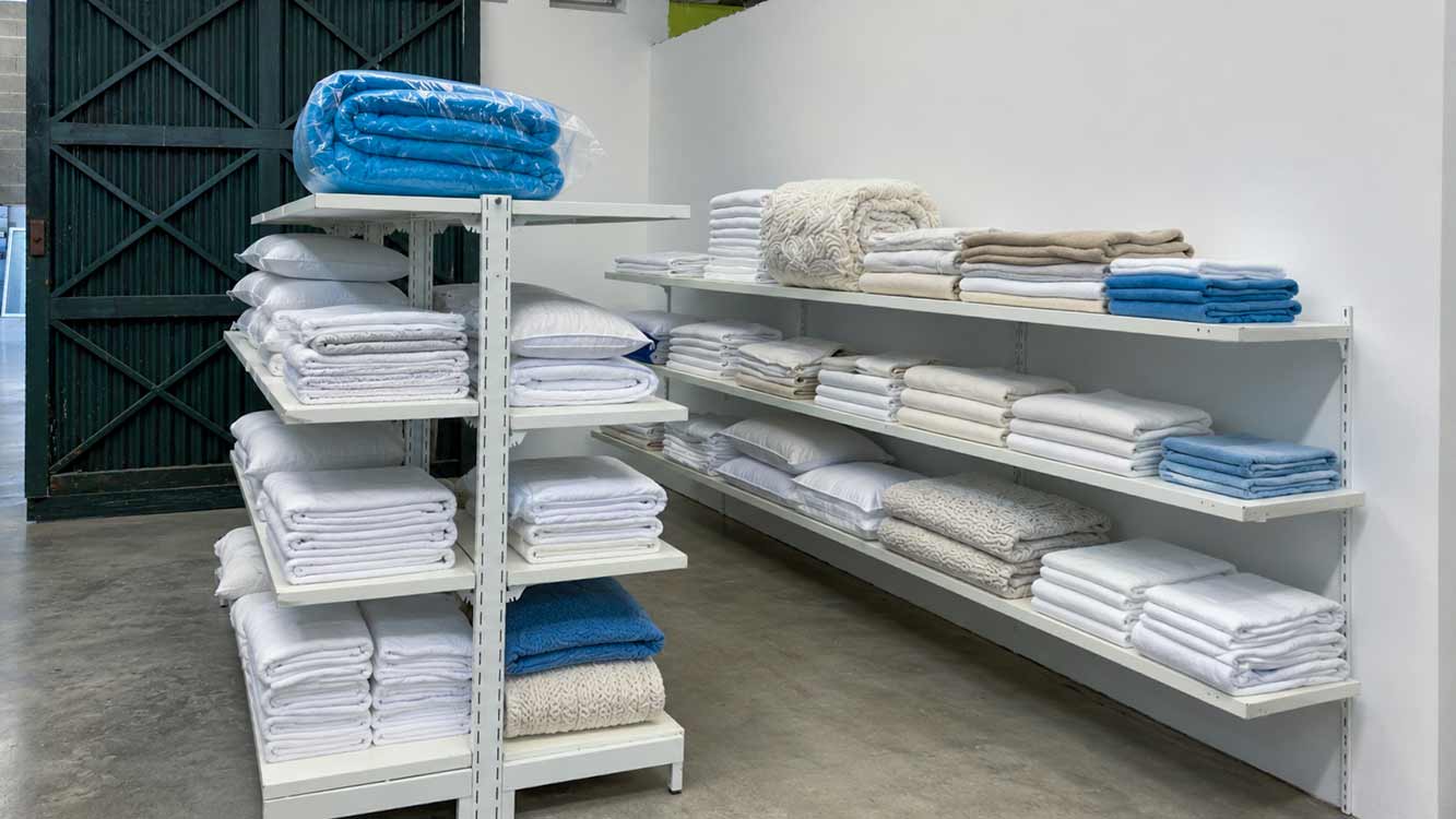 Estantes com roupa de cama dobrada, almofadas e toalhas arrumadas por prateleira.